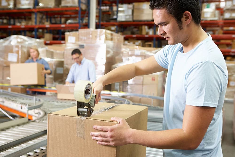 Warehouse worker sealing cardboard box with packing tape on conveyor belt